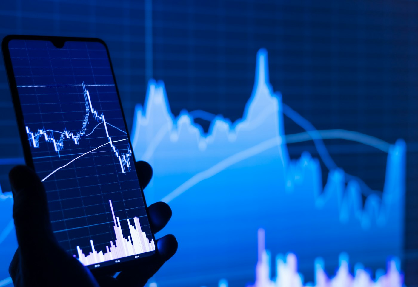 A phone showing a stock chart, held in front of a larger market chart
